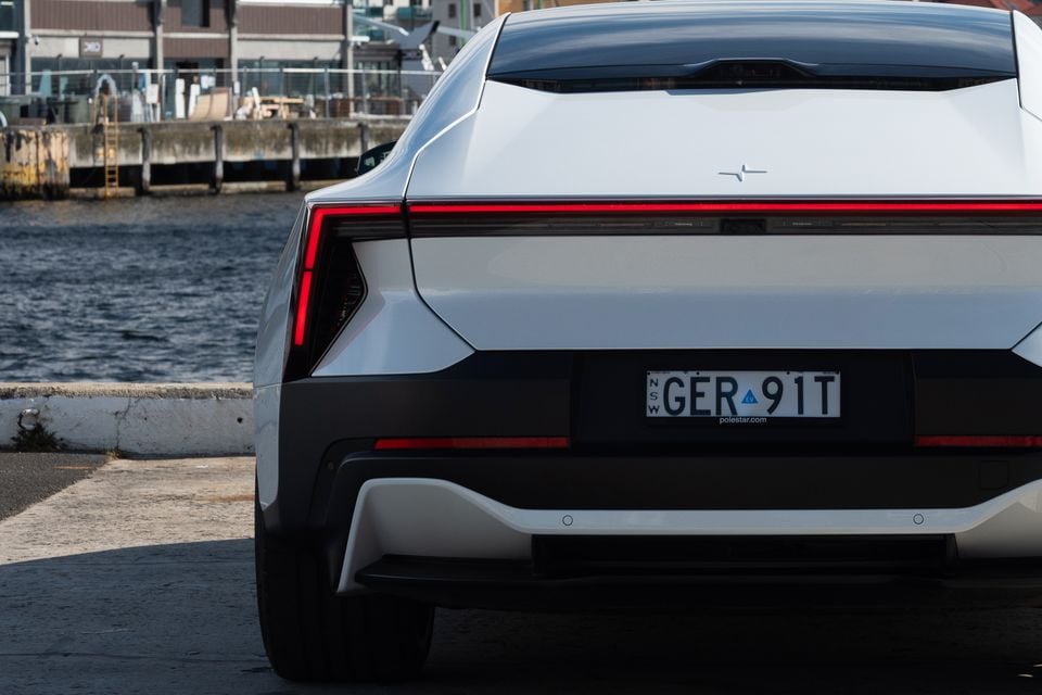 White 2027 Polestar 5 Performance seen from the rear, showing its full-width tail-light bar, parked beside a harbour wharf