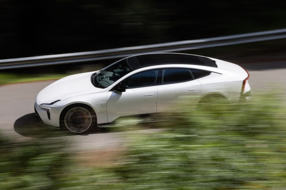 A white 2027 Polestar 5 Performance sedan driving along a road past a guardrail, seen from the side