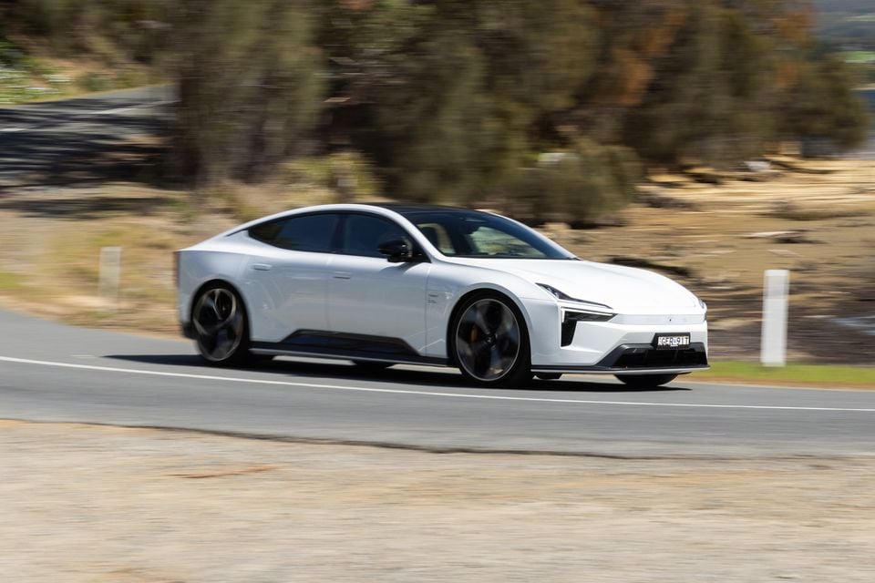 A white 2027 Polestar 5 Performance sedan driving along a rural road, seen from the front side