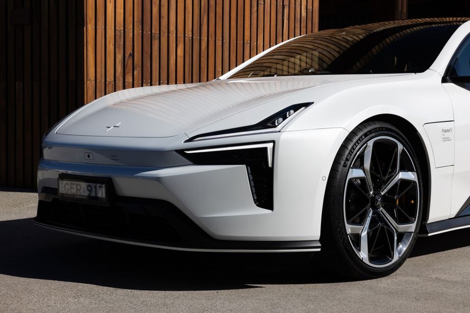Front end of a white 2027 Polestar 5 Performance showing its slim headlights, bonnet emblem and large alloy wheel, parked by a timber wall