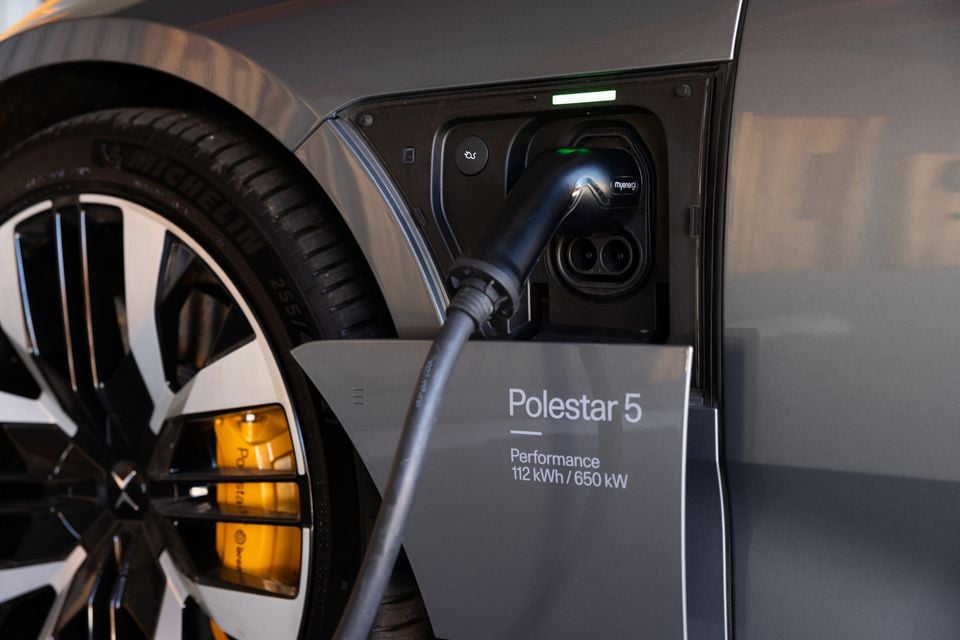 Close-up of a grey 2027 Polestar 5 Performance charging, with a cable plugged into its front charging port beside the wheel and yellow brake caliper