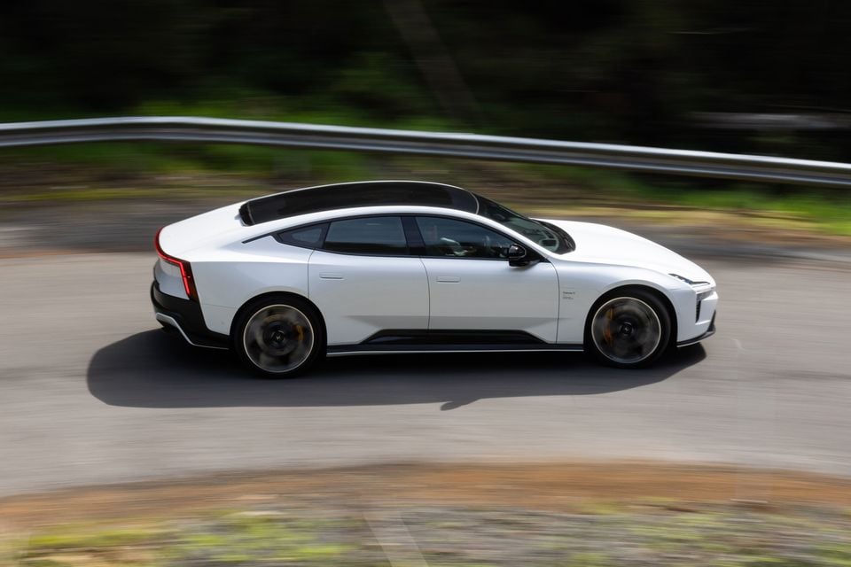 A white 2027 Polestar 5 Performance sedan driving along a winding road, seen side-on with a motion-blurred background