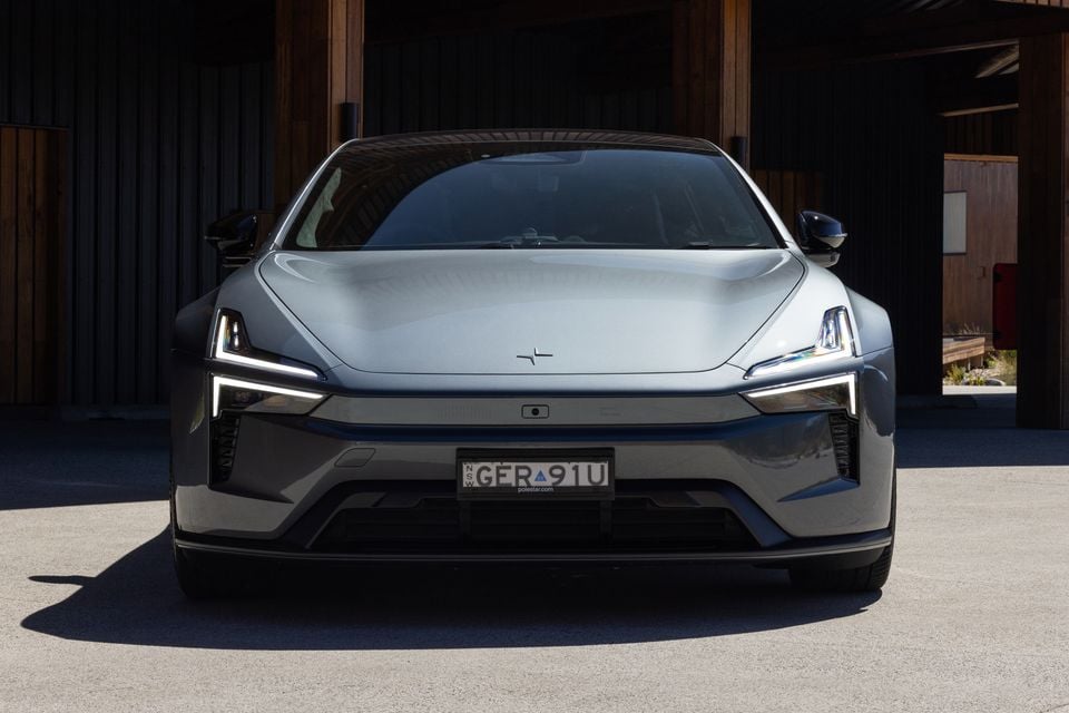 A grey 2027 Polestar 5 Performance seen from the front, parked in front of a dark timber building