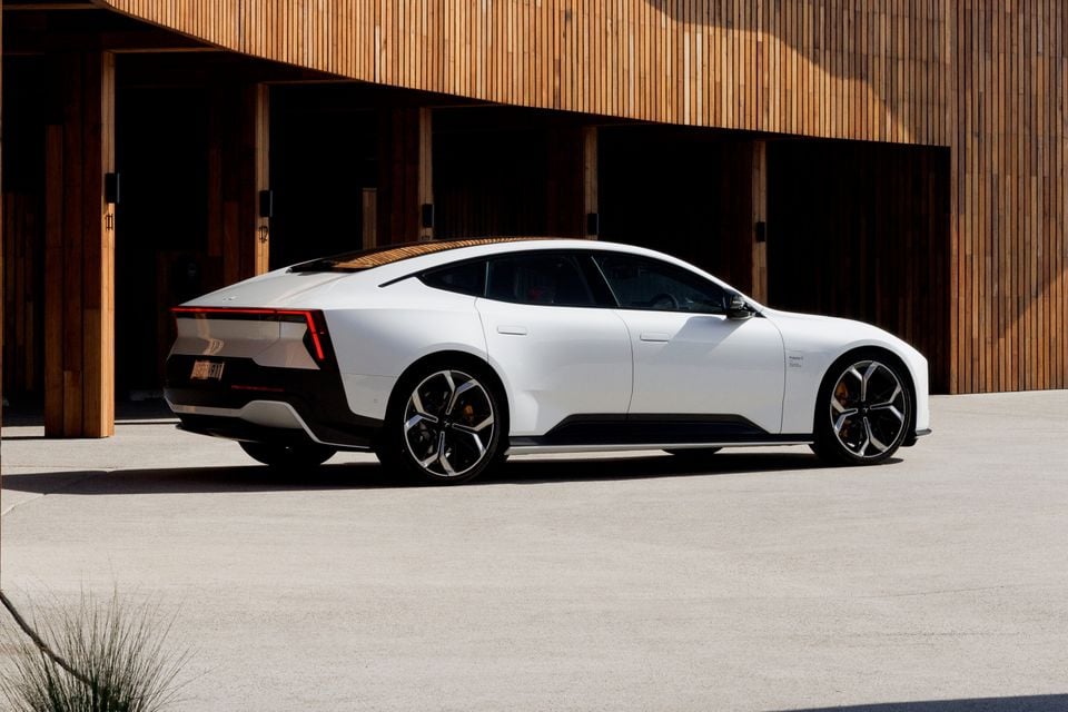 A white 2027 Polestar 5 Performance four-door grand tourer parked in front of a timber-clad building, seen from the rear side