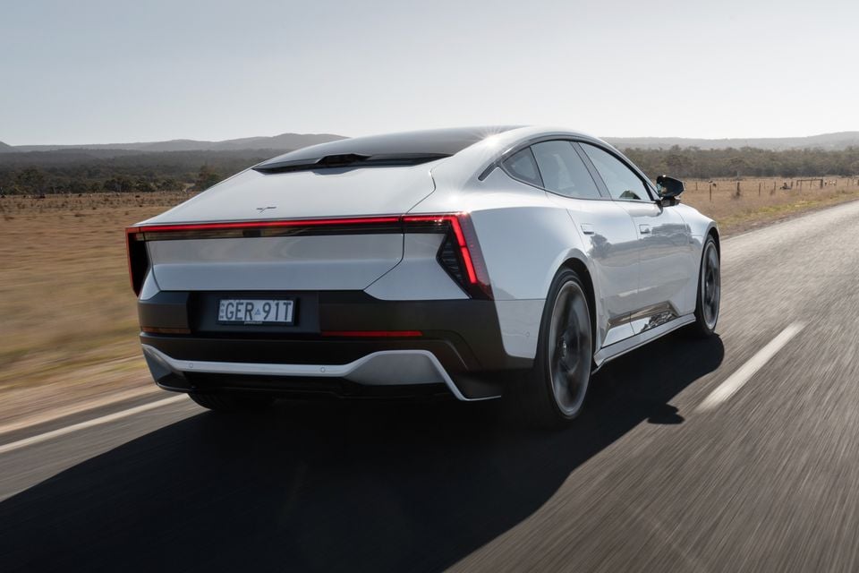 A white 2027 Polestar 5 Performance liftback driving on a country road, seen from the rear passenger side