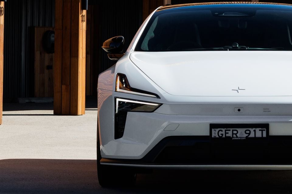 Front of a white 2027 Polestar 5 Performance, showing its slim headlight and bonnet badge, parked beside timber-clad posts
