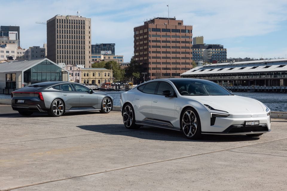 Um Polestar 5 Performance 2027 branco e cinza estacionado em um cais à beira-mar com edifícios urbanos atrás, um visto de frente e outro de trás