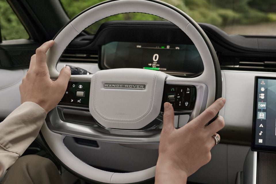 Hands on the Range Rover Electric steering wheel, showing the digital instrument cluster and touchscreen infotainment system inside the cabin.