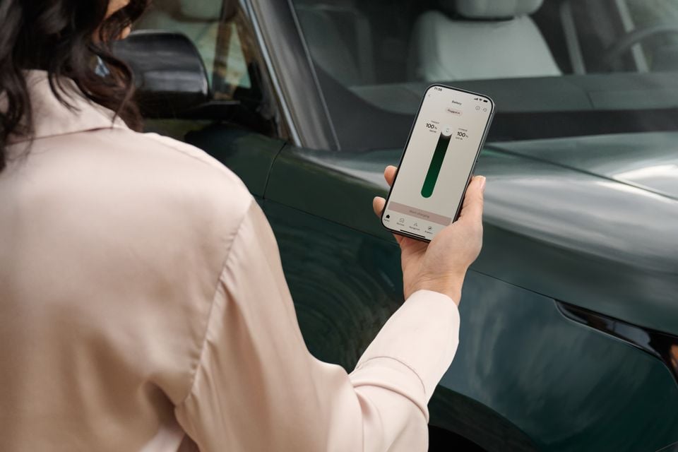 A person holding a smartphone displaying a battery charging app next to a dark green Range Rover Electric SUV