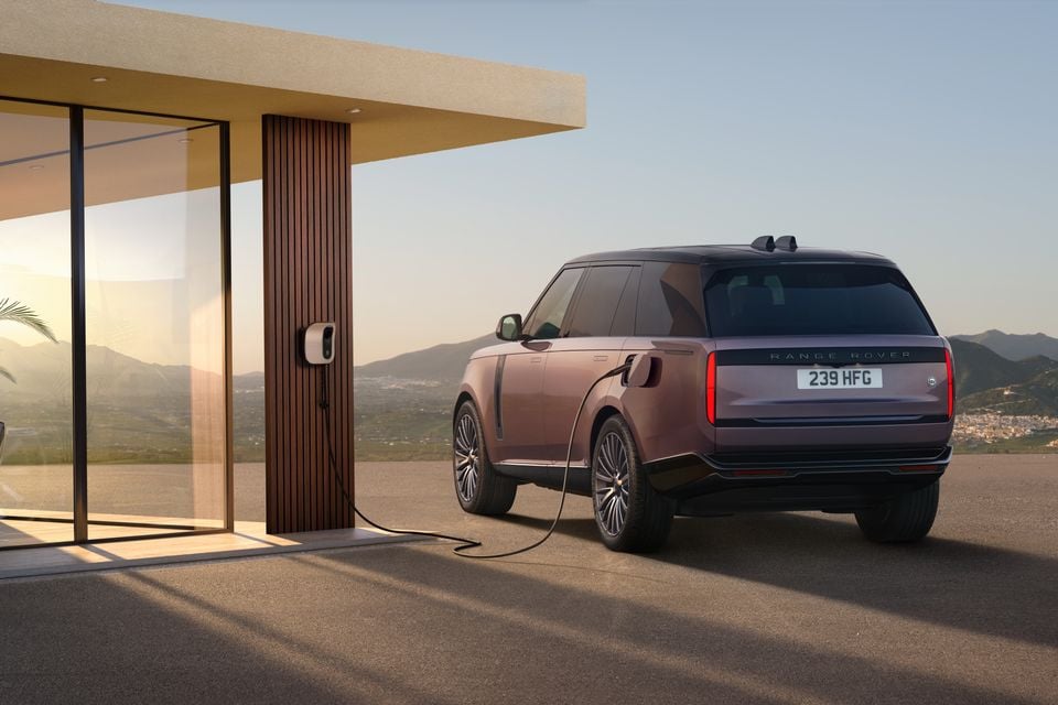 A Range Rover SUV seen from the rear three-quarter angle, plugged into a home EV wall charger outside a modern building with a scenic mountain backdrop.