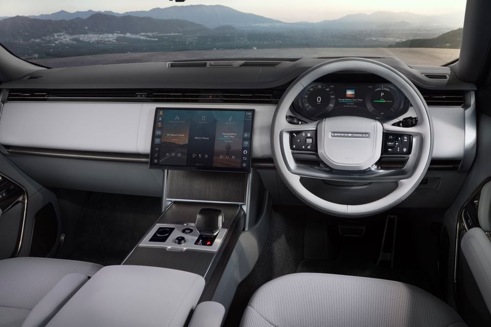Interior of the 2027 Range Rover Electric showing the minimalist dashboard, large central touchscreen infotainment display, white steering wheel, and light grey seats with a panoramic view through the windscreen