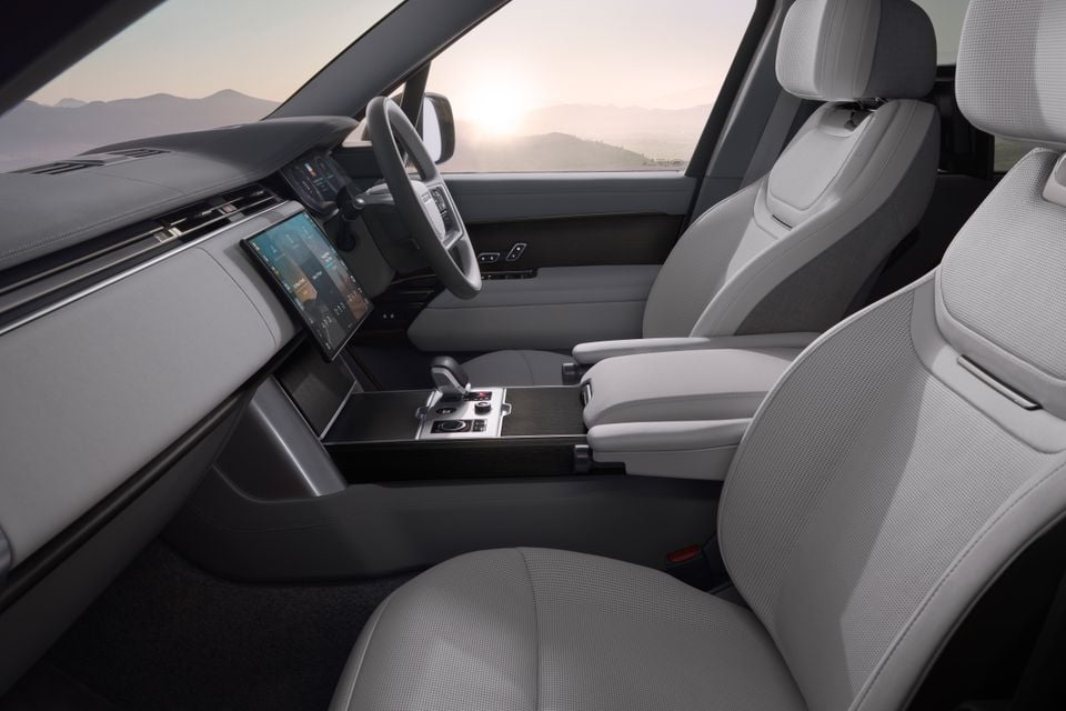 Interior of the 2027 Range Rover Electric showing light grey front seats, large touchscreen infotainment display, centre console, and steering wheel with a scenic mountain backdrop