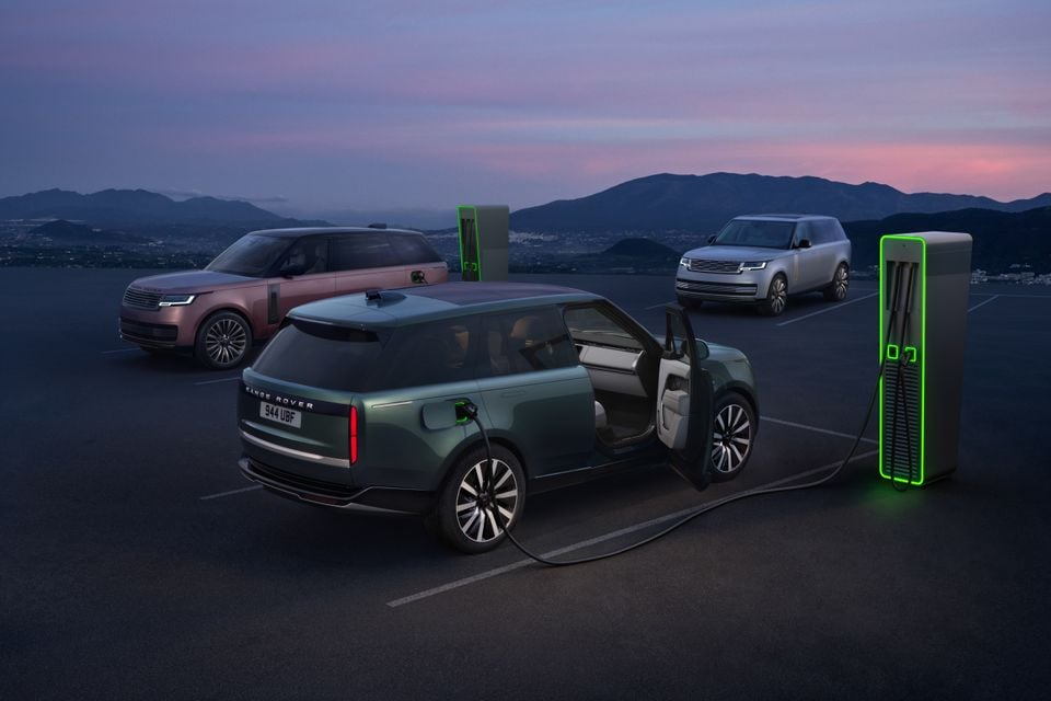 Three Range Rover Electric SUVs in different colours plugged into fast chargers at dusk, shown from the rear and side angles with mountains in the background