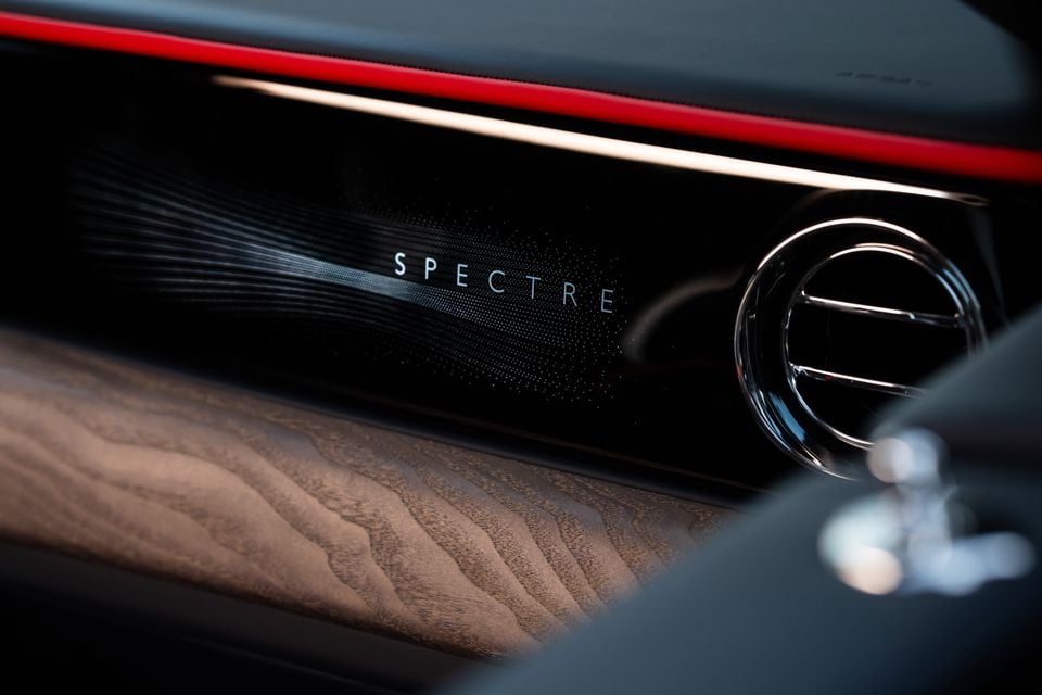 Illuminated Spectre lettering on the dashboard fascia of a 2027 Rolls-Royce Spectre, with a chrome air vent and open-pore wood trim