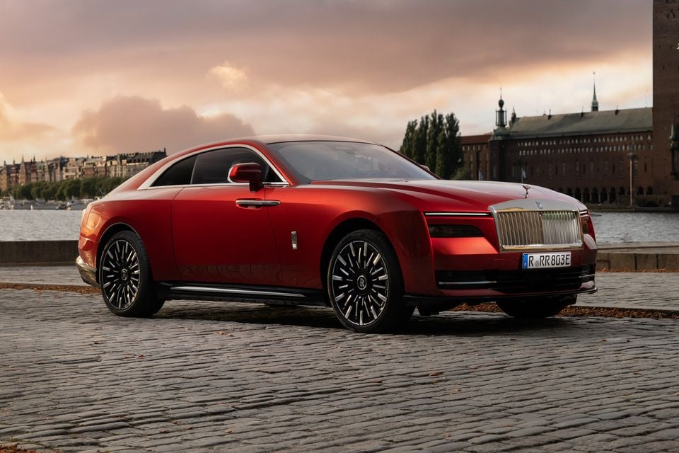 A red 2027 Rolls-Royce Spectre electric coupe seen from the front side, parked on a cobblestone waterfront at sunset