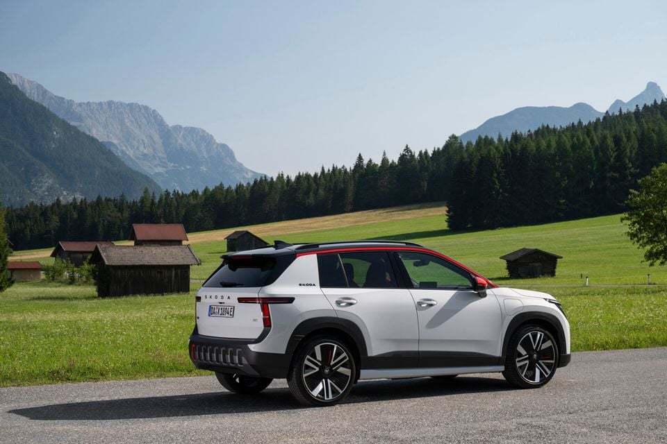 A white Skoda Epiq electric SUV seen from the rear three-quarter angle, parked on a road with alpine mountains and green meadows in the background