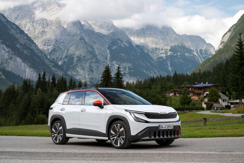 A white Skoda Epiq electric SUV seen from the front-side angle, parked in front of an alpine mountain landscape with red mirror caps and roof trim accents.