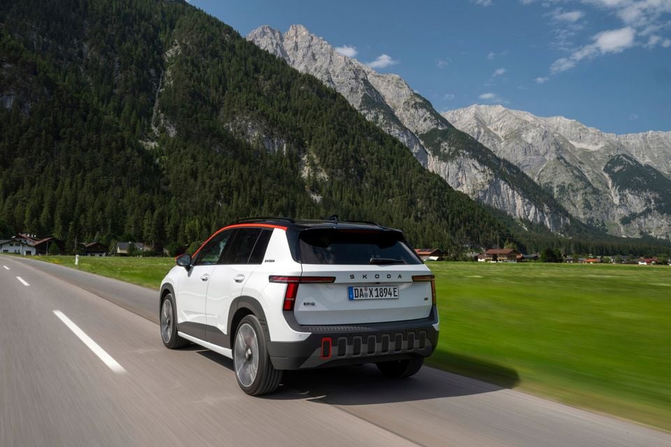 A white Skoda Epiq electric SUV driving along a mountain road, seen from the rear three-quarter angle with alpine scenery in the background.
