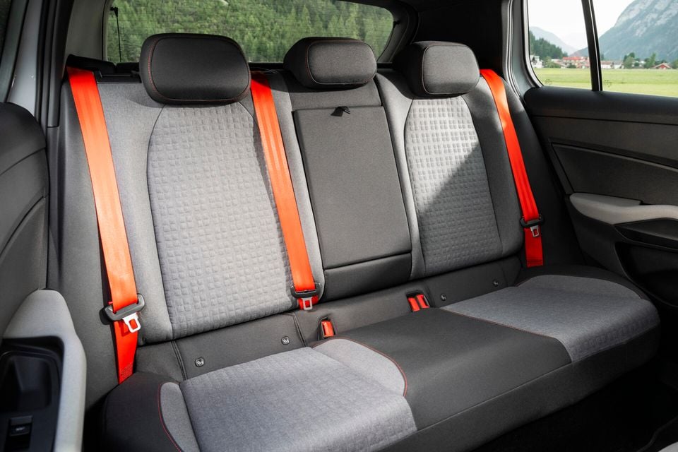 Rear passenger bench of a Skoda Epiq First Edition showing three seats with grey fabric upholstery, red stitching accents, and bright orange seatbelts, with an alpine landscape visible through the windows.