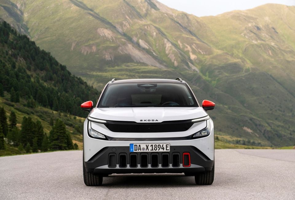 A white Skoda Epiq SUV seen from the front, with red mirror caps and accent details, parked on a road with green mountains in the background.