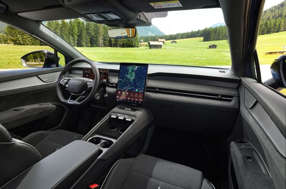 Interior of a Skoda Elroq showing the dashboard, large central touchscreen infotainment display, steering wheel, centre console and front seats, with a rural landscape visible through the windscreen.