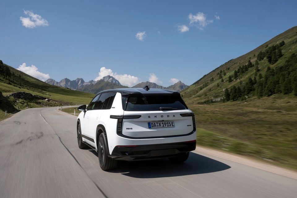 A white Skoda Peaq SUV seen from the rear while driving on a mountain road, with green hills and peaks in the background.