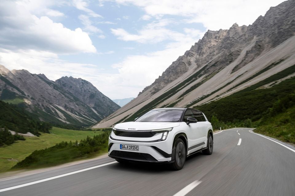 A white Skoda Peaq SUV driving along a mountain road with alpine scenery in the background, seen from the front