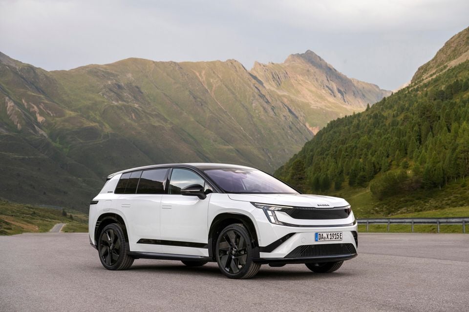 A white Skoda Peaq electric SUV seen from the front-side angle, parked on a mountain road with alpine scenery in the background.