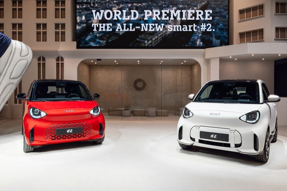 A red and a white 2027 Smart #2 city car displayed side by side under a World Premiere screen at a motor show stand