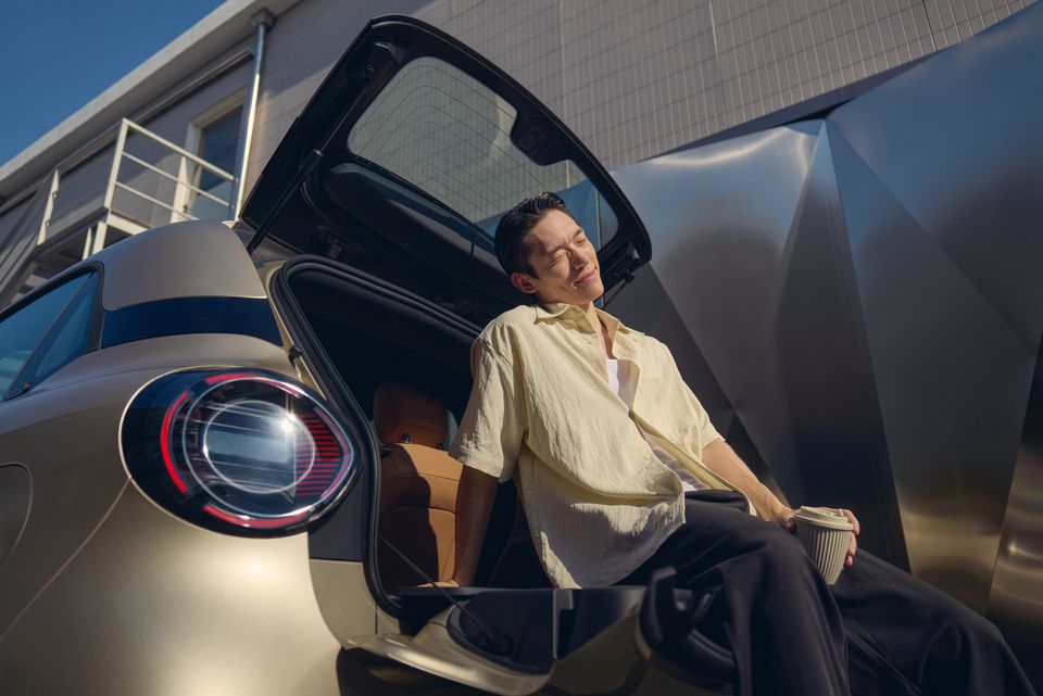 A champagne-coloured 2027 Smart #2 seen from the rear with its tailgate open, as a man sits in the boot holding a coffee cup