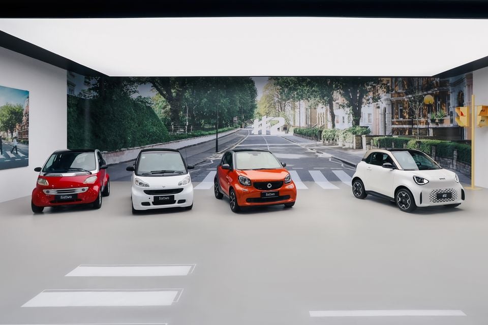 Four generations of Smart city cars, three Smart Fortwo models in red, white and orange beside a white Smart #2, displayed in a studio with a street backdrop