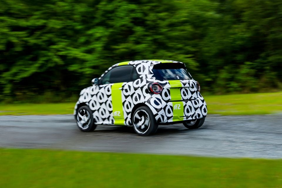 A camouflaged Smart #2 prototype hatchback seen from the rear three-quarter angle while driving on a road, wearing a black, white and lime green wrap