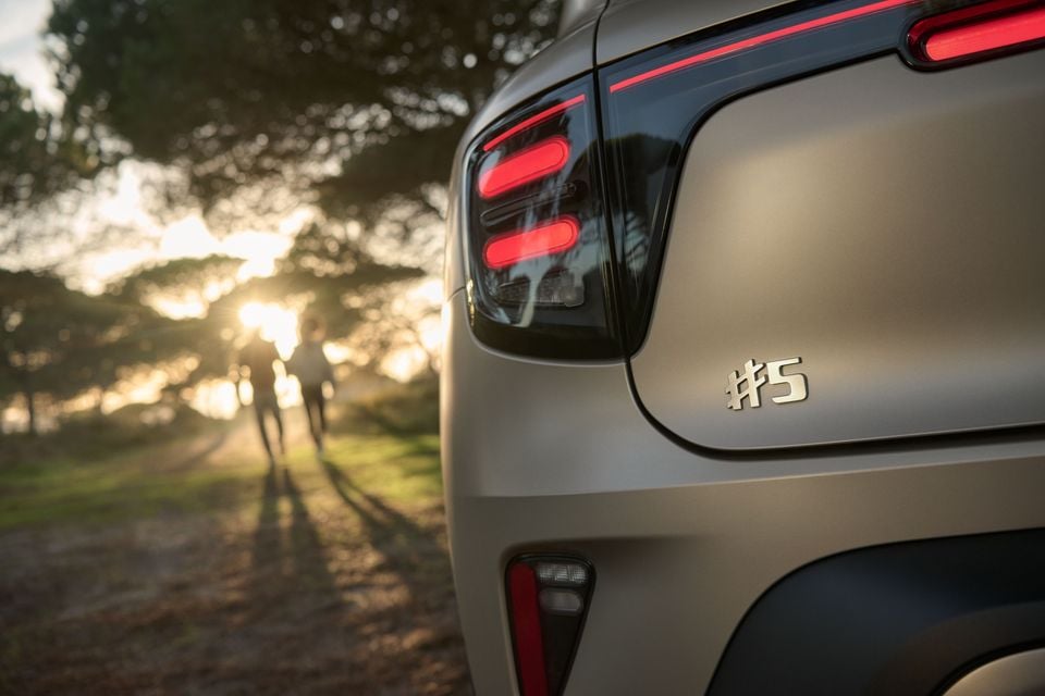 Close-up of the rear tail light and #5 badge on a Smart #5 SUV, with a golden sunset and people walking in the background