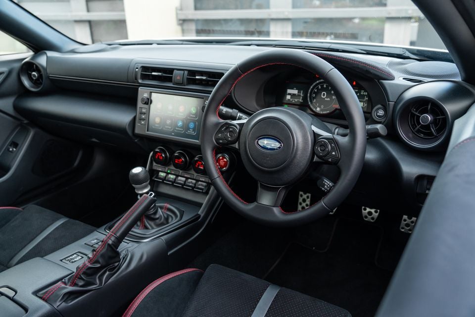 Interior of a Subaru BRZ showing the steering wheel with red stitching, manual gear shifter, touchscreen infotainment system, and sport pedals from the driver's side perspective