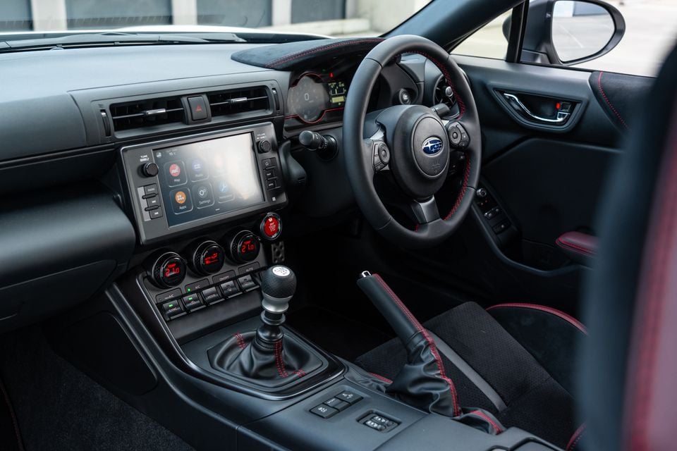 Interior of a Subaru BRZ showing the dashboard, manual gear shifter, handbrake, steering wheel with Subaru badge, and infotainment touchscreen with red accent stitching throughout