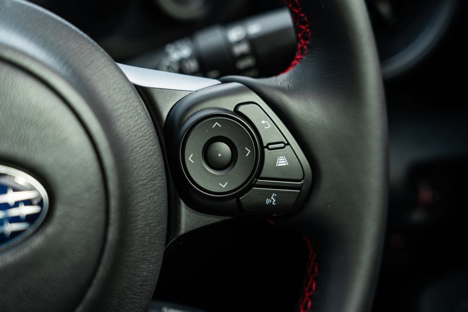 Close-up of the steering wheel controls on a Subaru BRZ, showing a directional pad and driver assist buttons with red stitching detail visible on the leather rim