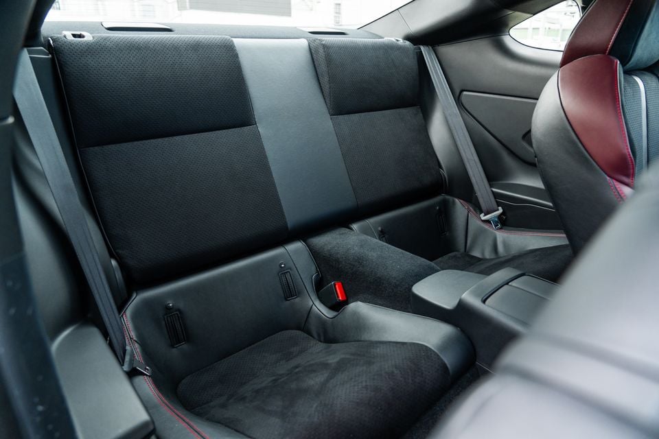Rear seats of a Subaru BRZ coupe, showing black leather and suede upholstery with red stitching accents and seatbelts visible