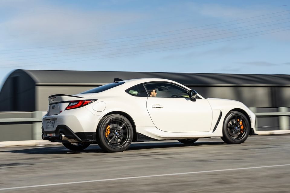 A white Subaru BRZ tS coupe seen from the rear three-quarter angle while driving on a road, featuring black wheels with yellow brake calipers and a rear spoiler