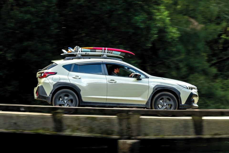A white Subaru Crosstrek SUV with surfboards on its roof rack driving across a bridge, seen from the side