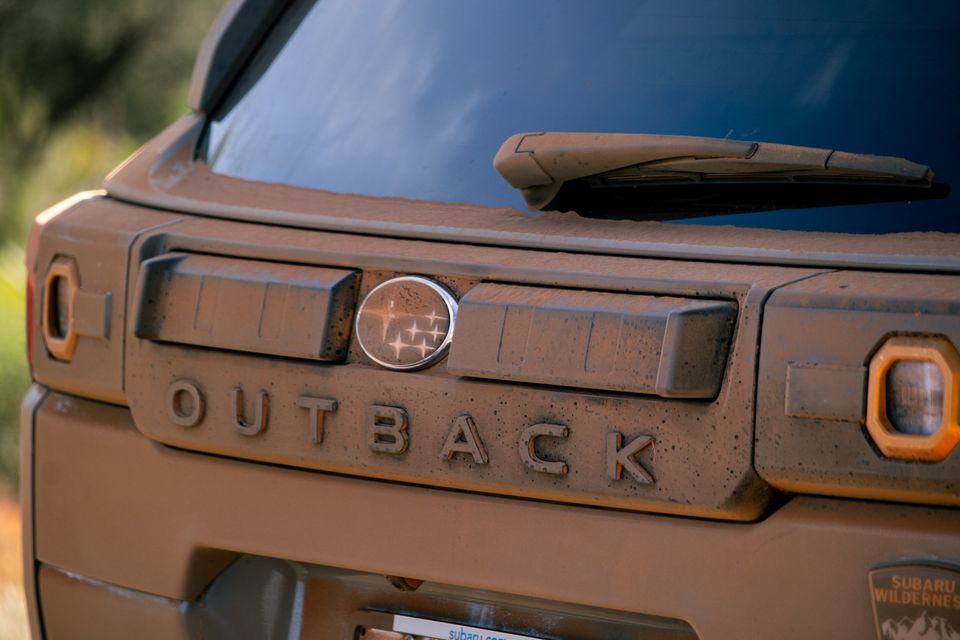 Close-up of the mud-covered rear tailgate of a Subaru Outback Wilderness, showing the Subaru badge and Outback lettering caked in dirt after off-road driving.