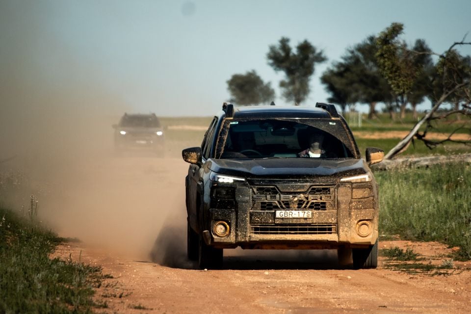 A mud-covered Subaru Outback Wilderness driving on a dusty dirt road in the Australian outback, seen from the front, with a second vehicle following behind in the dust