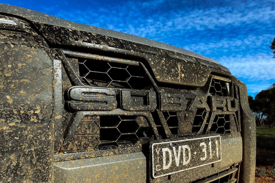Close-up of a mud-covered Subaru front grille with the Subaru badge visible, photographed outdoors under a blue sky.