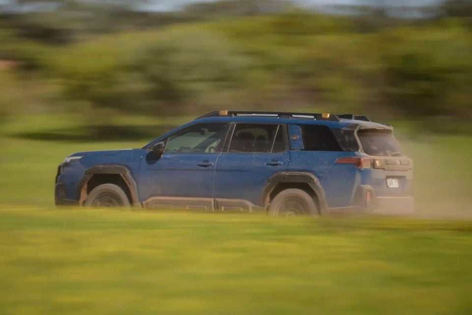 A blue Subaru Outback Wilderness SUV seen from the side, driving across a grassy field and kicking up dust