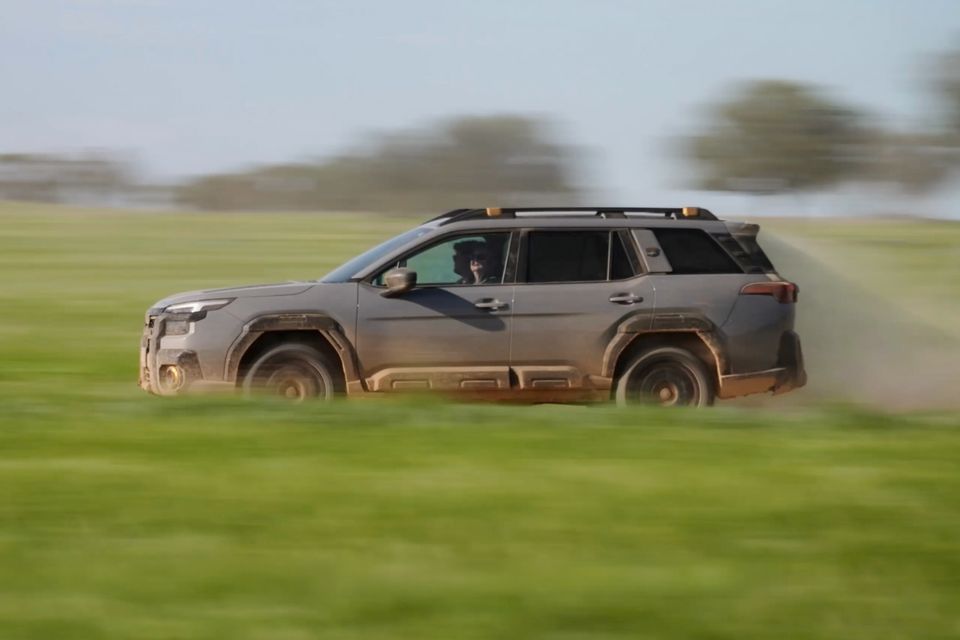 A grey Subaru Outback Wilderness SUV driving at speed across a grassy field, seen from the side, covered in mud and dust