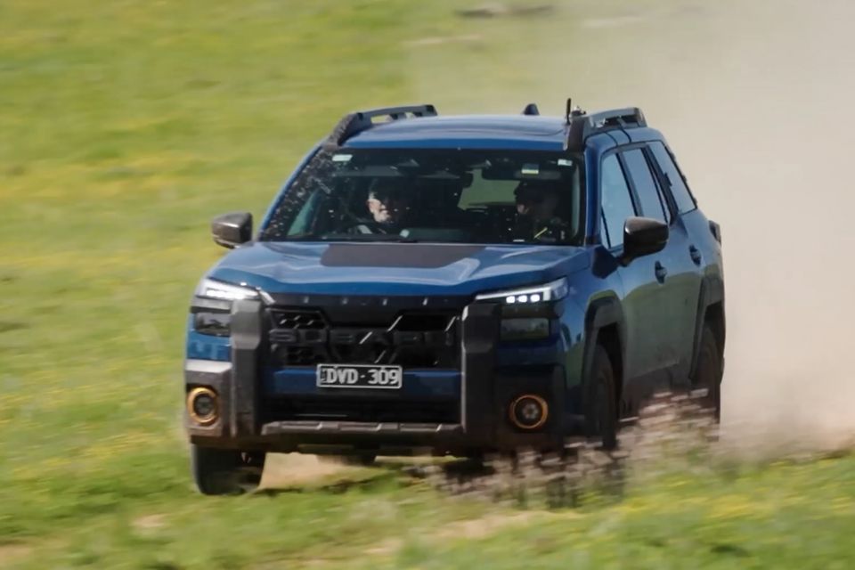 A blue Subaru Outback Wilderness SUV driving off-road across a grassy field, kicking up dust, seen from the front