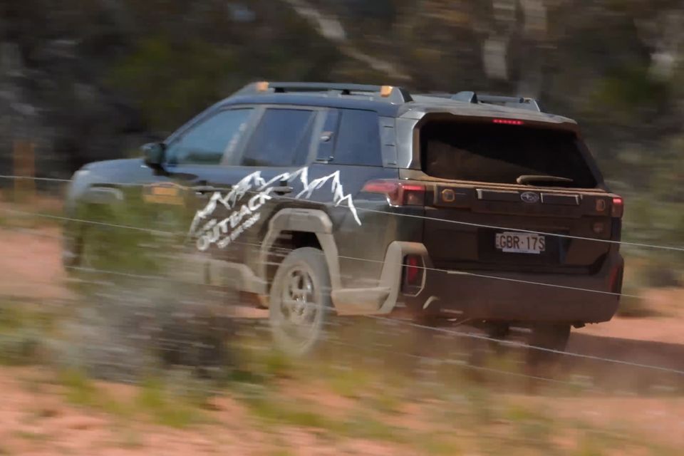 A Subaru Outback Wilderness SUV driving along a dirt track, seen from the rear three-quarter angle, with an adventure-themed wrap on the side panel