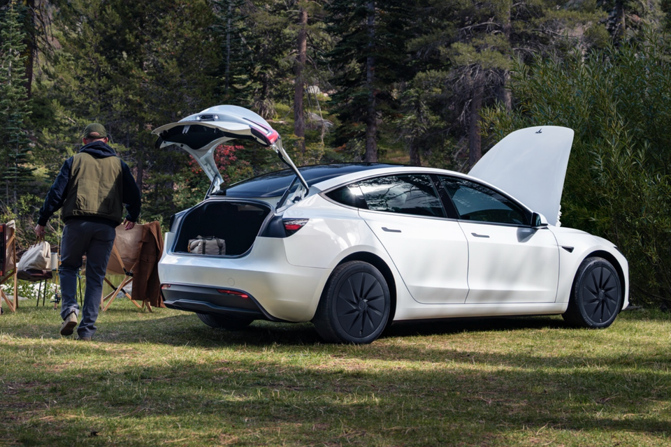 A white Tesla Model Y with its boot and bonnet open, parked on grass in a forest while a man carries camping gear nearby.