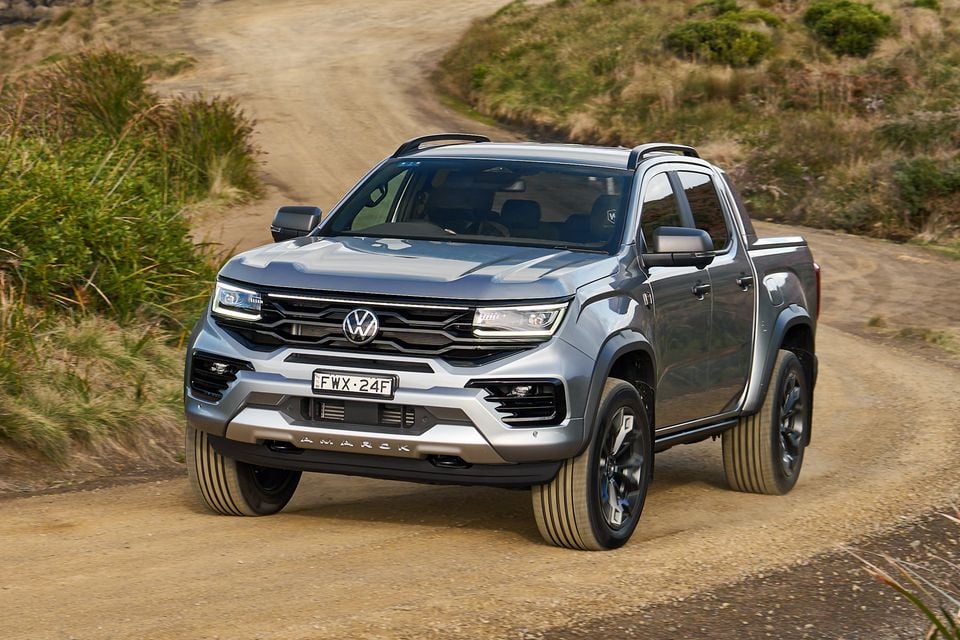 A silver Volkswagen Amarok dual-cab ute driving on a dirt track in an outdoor rural setting, seen from the front three-quarter angle
