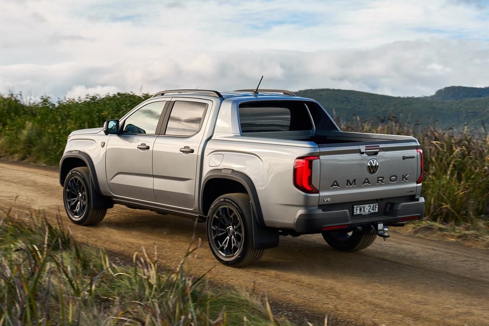 A silver Volkswagen Amarok V6 dual-cab ute seen from the rear three-quarter angle, driving along a dirt road with mountains in the background