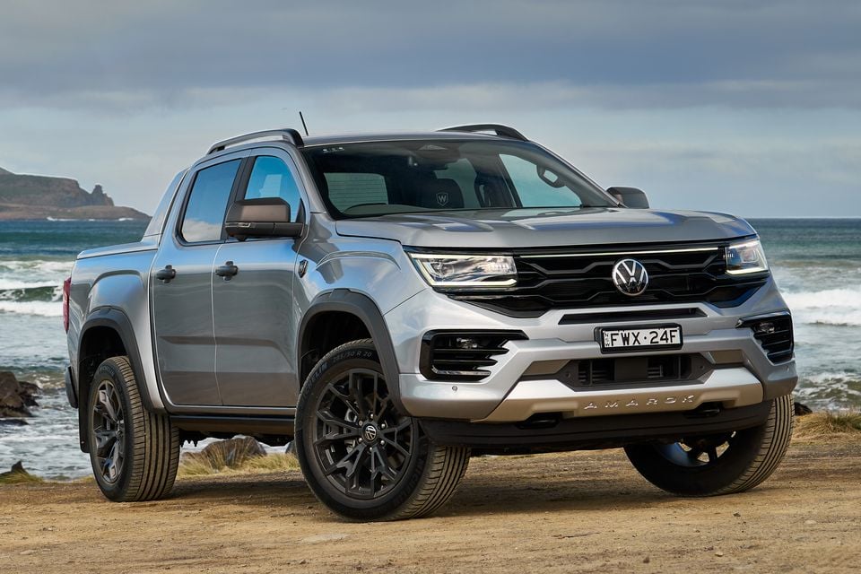 A silver Volkswagen Amarok dual-cab ute photographed from the front-side angle on a coastal dirt area with ocean waves in the background